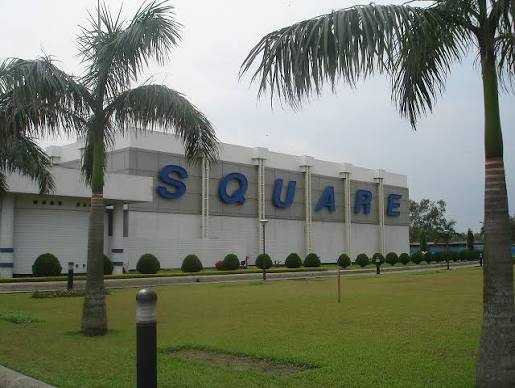 A white factory building with large blue letters spelling Square, behind palm trees and a lawn.